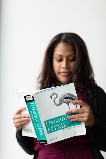 The image shows a woman with long dark hair wearing a black blazer and a maroon top, standing indoors against a plain white background. She is attentively examining a gardening book titled 'Dynamic HT