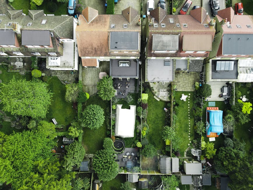 An aerial view of a residential neighborhood showing a row of terraced houses with varied rooftops, including tiled and flat roofs. Behind the houses are garden areas featuring lush green lawns, matur